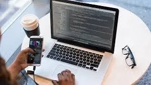 Person holding a phone beside a laptop displaying code on a round table